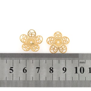 May include: Two gold-colored metal flower-shaped bead caps, each measuring approximately 7 mm in diameter, are shown on a ruler. The bead caps have a delicate, intricate design.