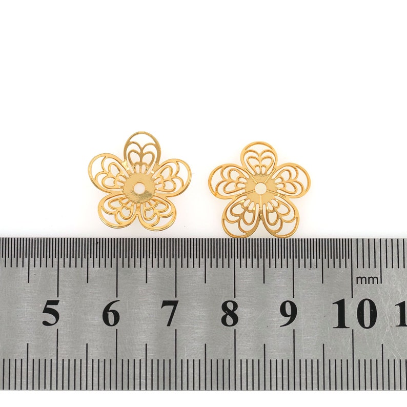 May include: Two gold-colored metal flower-shaped bead caps, each measuring approximately 7 mm in diameter, are shown on a ruler. The bead caps have a delicate, intricate design.