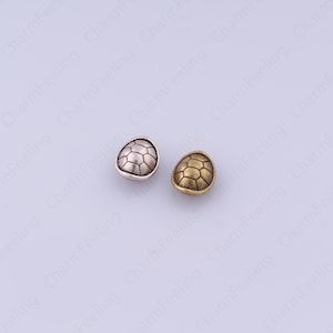 May include: Two metal turtle shell shaped beads. One bead is silver-colored, and the other is gold-colored. The beads have a geometric pattern on the surface, resembling a turtle shell.