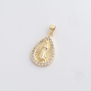 May include: Gold-colored pendant in a teardrop shape, featuring a religious figure in the center. The pendant is surrounded by a border of small, clear gemstones. The bail is also gold-colored.