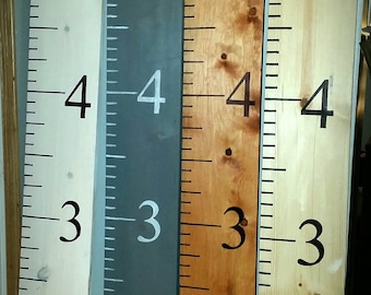 May include: Four wooden growth charts with black numbers and markings. The charts are painted in white, grey, light cherry, and natural wood finishes. The bottom of each chart is labelled with the corresponding wood finish.