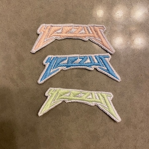 May include: Three embroidered patches in pastel colors: peach, light blue, and pale green. Each patch features a stylized, angular design with white stitching. The patches are arranged on a neutral-toned surface.