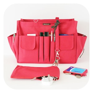 Op de afbeelding: Een rode tote bag met meerdere zakken en compartimenten, waaronder een afneembare etui en een kleine portemonnee. De tas heeft een handvat en een schouderriem. De tas is gevuld met pennen, sleutels en andere accessoires.