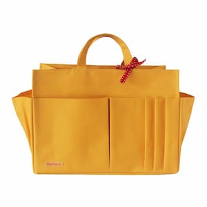 Può includere: Una borsa organizer rettangolare color giallo senape con due manici superiori, dotata di molteplici tasche esterne di varie larghezze sul davanti, soffietti laterali e un piccolo nastro rosso a motivi annodato a un manico. Un'etichetta è in basso a sinistra.