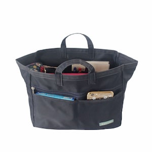 Peut inclure: Un sac fourre-tout noir avec plusieurs poches et un compartiment zippé. Le sac est rempli de divers objets, dont un téléphone, un livre et des stylos. Le sac porte une étiquette indiquant "Myllute".
