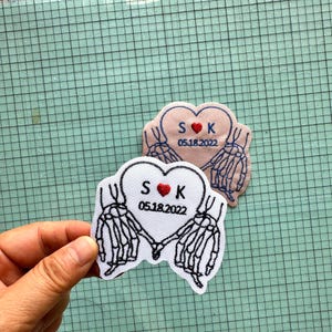Könnte beinhalten: Zwei gestickte Aufnäher mit einem Herzdesign und sich haltenden Skeletthänden. Die Aufnäher haben die Initialen "S" und "K" mit einem roten Herz und dem Datum "05.18.2022". Ein Aufnäher ist weiß, der andere hellrosa.