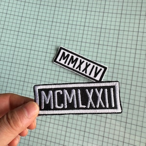 Könnte beinhalten: Zwei weiße Stoffpatches mit schwarzen gestickten römischen Ziffern. Der obere Patch lautet "MMXXIV" und der untere Patch lautet "MCM LXXII".