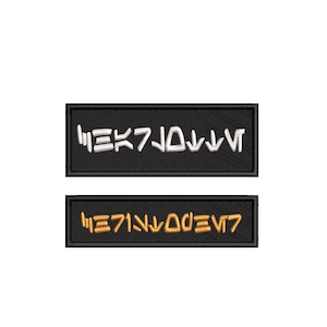 Könnte beinhalten: Zwei schwarze rechteckige Patches mit weiß und gold gesticktem Text. Der obere Patch hat weißen Text, der "HEK7J0JVV" lautet. Der untere Patch hat goldenen Text, der "HE71NVOOEV7" lautet.