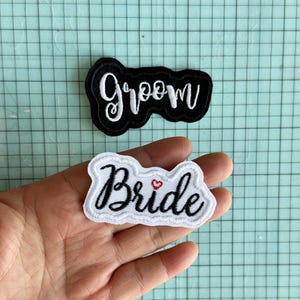 May include: Two white fabric patches with black embroidered lettering. One patch says "Groom" and the other says "Bride" with a small red heart.