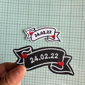 Könnte beinhalten: Zwei gestickte Patches mit einem Banner-Design. Der obere Patch ist weiß mit schwarzen Nähten und der untere Patch ist schwarz mit weißen Nähten. Beide Patches haben das Datum "24.02.22" und rote Herzen.