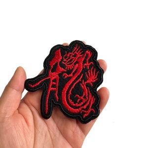 May include: Embroidered patch featuring a red dragon design on a black background. The dragon is intricately detailed with a stylized Chinese character. The patch has a black border and is likely for clothing or accessories.