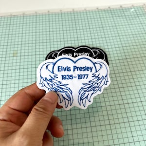 Op de afbeelding: Een witte patch met blauw geborduurde tekst en vleugelontwerp. De tekst luidt "Elvis Presley 1935-1977". Een zwarte patch met de tekst "Elvis Presley" is zichtbaar op de achtergrond. De patches zijn waarschijnlijk voor kleding of accessoires.