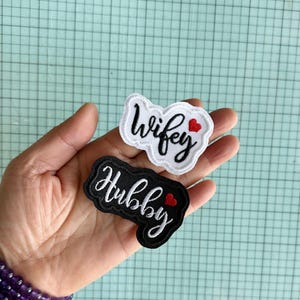 May include: Two embroidered patches, one white with the word "wifey" and a red heart, and one black with the word "hubby" and a red heart.