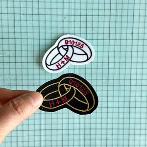 May include: Two embroidered patches featuring interlocking wedding rings. One patch is black with gold stitching, the other is white with black stitching. Both patches have the date "010125" and the initials "H+M" embroidered in red.