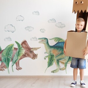 May include: A young child wearing a cardboard dinosaur costume, holding a large cardboard box. The background features a wall mural with watercolor-style dinosaur illustrations and fluffy clouds.