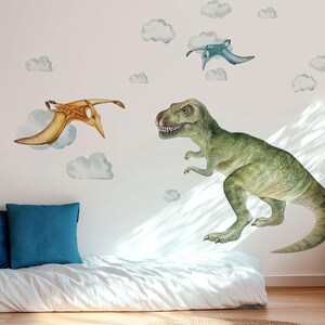 May include: Watercolor dinosaur wall decals featuring a green T-Rex and two pterodactyls in flight, with fluffy white clouds. The decals are applied to a white wall in a child's bedroom, above a low bed with blue pillows.