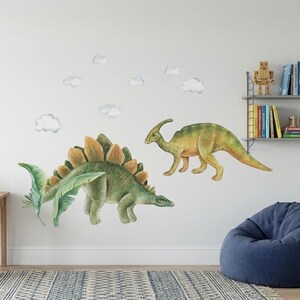 May include: Two dinosaur wall decals, a green Stegosaurus with orange plates and a yellow and green Parasaurolophus, are displayed on a white wall. Several white cloud decals are also present. A desk with a solar system poster and a globe are in the background.