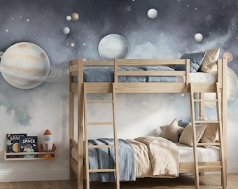May include: A light-colored wooden bunk bed sits against a wall mural featuring a dark blue and grey outer space scene with various planets, stars, and nebulae. A small wall-mounted shelf holds two books titled Space and a small toy rocket ship.