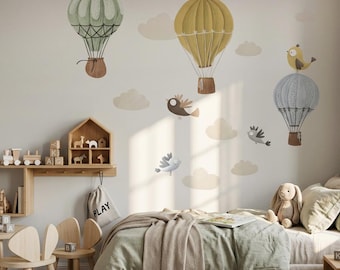 May include: A wall decal set featuring three hot air balloons in sage green, mustard yellow, and light blue, each with a small bird perched on top. The design includes several beige clouds and two small flying birds, creating a whimsical scene.