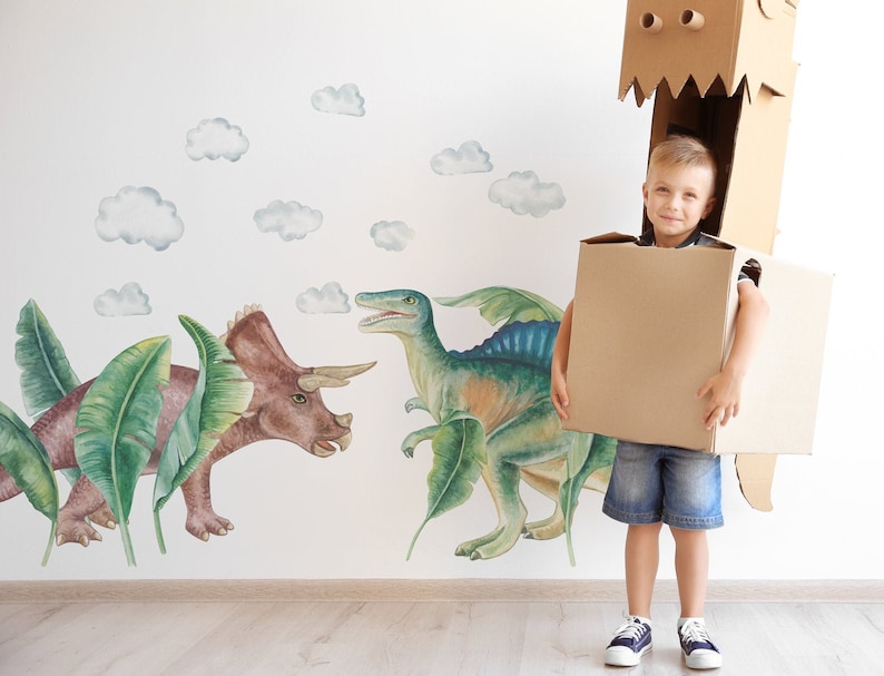 May include: A young child wearing a cardboard dinosaur costume, holding a large cardboard box. The background features a wall mural with watercolor-style dinosaur illustrations and fluffy clouds.