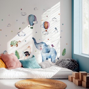 K&ouml;nnte beinhalten: Ein Kinderzimmer mit Aquarell-Tierwandaufklebern, darunter ein Elefant, eine Schildkr&ouml;te und ein Strau&szlig;, sowie Hei&szlig;luftballons und Wolken. Der Raum ist mit bunten Kissen und einem runden Teppich ausgestattet und schafft eine verspielte und einladende Atmosph&auml;re.