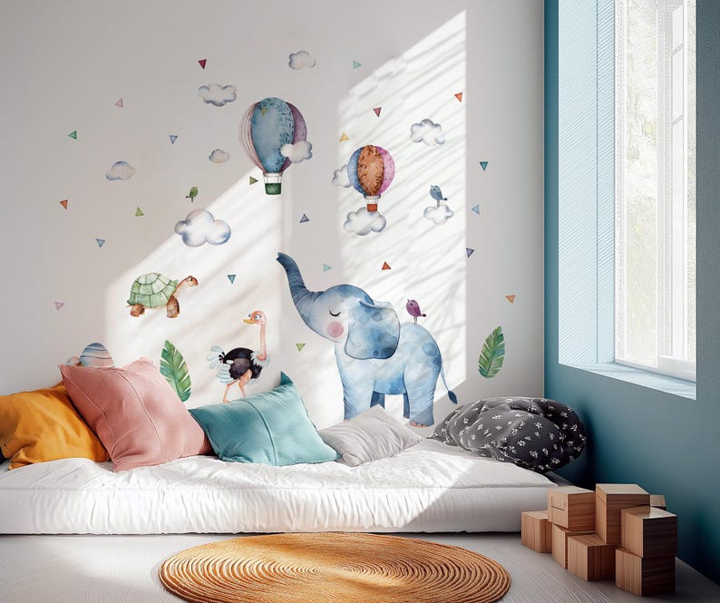K&ouml;nnte beinhalten: Ein Kinderzimmer mit Aquarell-Tierwandaufklebern, darunter ein Elefant, eine Schildkr&ouml;te und ein Strau&szlig;, sowie Hei&szlig;luftballons und Wolken. Der Raum ist mit bunten Kissen und einem runden Teppich ausgestattet und schafft eine verspielte und einladende Atmosph&auml;re.
