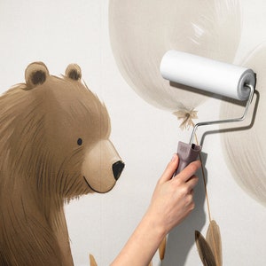 May include: A wall mural featuring a brown bear illustration next to light beige balloons. A white paint roller is being used to apply the mural to the wall surface. The bear has dark brown fur, a tan muzzle, and a black nose, while the balloons are depicted with soft, brush-like strokes.