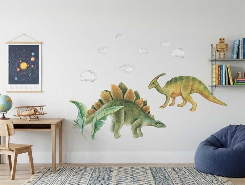 May include: Two dinosaur wall decals, a green Stegosaurus with orange plates and a yellow and green Parasaurolophus, are displayed on a white wall. Several white cloud decals are also present. A desk with a solar system poster and a globe are in the background.