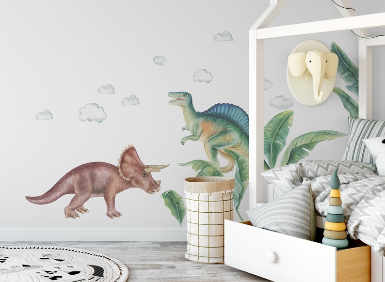 May include: Watercolor dinosaur wall decals featuring a Triceratops and a Spinosaurus in shades of brown, green, and blue. The wall art includes cloud accents, creating a playful, prehistoric theme for a child's room.