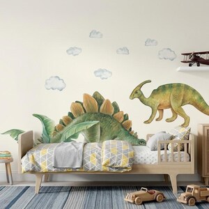 May include: A child's bedroom with a dinosaur theme. The room features a wooden bed with a yellow and gray patterned blanket, dinosaur wall decals, and a wooden toy truck and car. A lamp with dinosaur illustrations sits on a wooden cabinet.