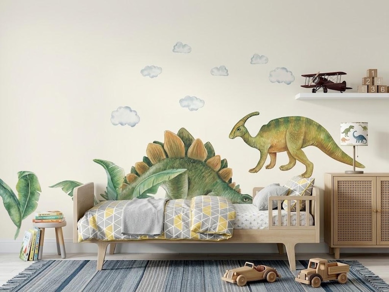 May include: A child's bedroom with a dinosaur theme. The room features a wooden bed with a yellow and gray patterned blanket, dinosaur wall decals, and a wooden toy truck and car. A lamp with dinosaur illustrations sits on a wooden cabinet.