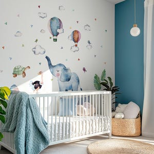 K&ouml;nnte beinhalten: Ein Kinderzimmer mit einem wei&szlig;en Kinderbett und einer blauen Decke. Die Wand ist mit Aquarellillustrationen von Hei&szlig;luftballons, Wolken, einer Schildkr&ouml;te, einem Strau&szlig; und einem blauen Elefanten dekoriert. Ein Weidenkorb und ein runder Teppich sind ebenfalls zu sehen.