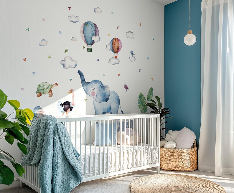 K&ouml;nnte beinhalten: Ein Kinderzimmer mit einem wei&szlig;en Kinderbett und einer blauen Decke. Die Wand ist mit Aquarellillustrationen von Hei&szlig;luftballons, Wolken, einer Schildkr&ouml;te, einem Strau&szlig; und einem blauen Elefanten dekoriert. Ein Weidenkorb und ein runder Teppich sind ebenfalls zu sehen.