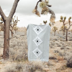 Peut inclure: Une couverture en tissu gris et blanc à motifs géométriques avec des accents noirs est suspendue à une branche d'arbre dans un paysage désertique.