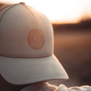 Puede incluir: Gorra de camionero beige con un parche circular con la palabra "WILD" y flechas. La gorra tiene una visera curva y una parte trasera de malla. La gorra es de color neutro y es adecuada para actividades al aire libre.