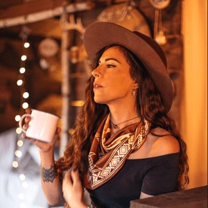 Peut inclure: Une femme portant un chapeau de cow-boy marron, un bandana marron et blanc, un haut noir à épaules dénudées, un jean bleu et une ceinture marron tient une tasse en émail blanche.