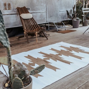 Peut inclure: Un tapis blanc avec un motif géométrique marron est posé sur une terrasse en bois. Une chaise en bois avec un coussin crème est près d'une caravane argentée vintage. Des cactus en pot sont au premier plan.
