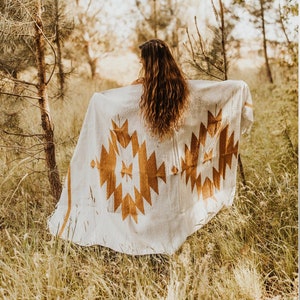 May include: A woman with long brown hair stands in a field of tall grass, holding a white blanket with a brown geometric pattern. The blanket has a southwestern design.