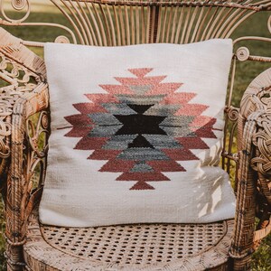 Peut inclure: Un coussin blanc avec un motif géométrique dans les tons rose, gris et noir posé sur un fauteuil en rotin en forme de paon.