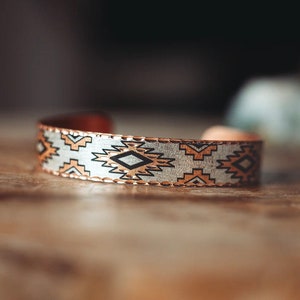 Peut inclure: Un bracelet en cuivre avec un motif géométrique en noir, blanc et orange. Le bracelet est posé sur une surface en bois.