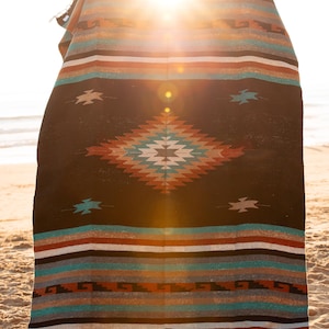 Puede incluir: Una manta tejida con un diseño del suroeste, con un patrón de diamante central en tonos de rojo, turquesa y blanco sobre un fondo negro. Rayas horizontales en varios colores en la parte superior e inferior. La manta se muestra en una playa de arena.