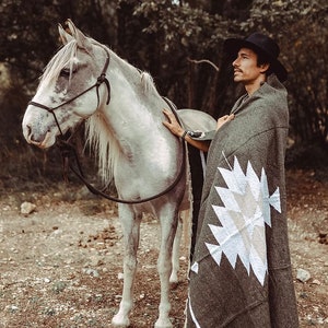 May include: A man wearing a black hat and a grey blanket with a white geometric pattern stands next to a white horse with a black halter.