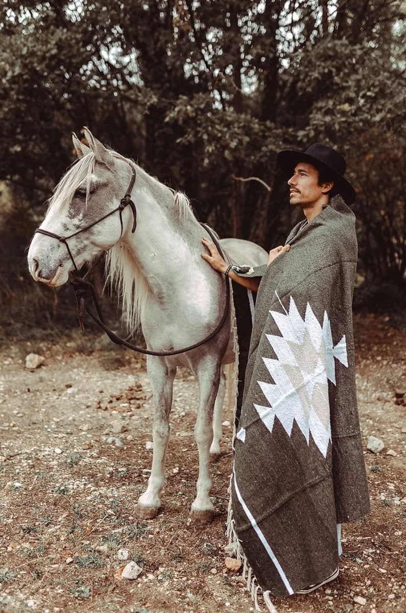 May include: A man wearing a black hat and a grey blanket with a white geometric pattern stands next to a white horse with a black halter.