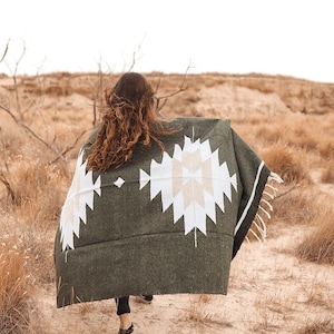 May include: A person walks across a desert landscape wearing a green and white patterned blanket. The blanket has a geometric design with white and beige accents.