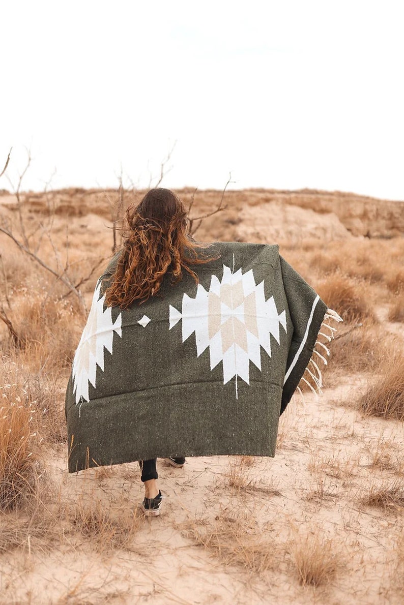 May include: A person walks across a desert landscape wearing a green and white patterned blanket. The blanket has a geometric design with white and beige accents.