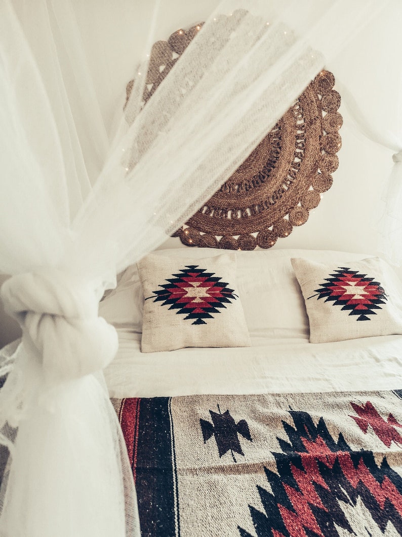 Pu&ograve; includere: Un letto bianco con un baldacchino bianco e una testiera in juta intrecciata. Il letto ha un piumone bianco e due cuscini con un motivo geometrico rosso e nero. Una coperta a motivi rosso, nero e bianco si trova ai piedi del letto.