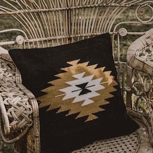 K&ouml;nnte beinhalten: Ein schwarzes Kissen mit goldfarbenem Muster liegt auf einem wei&szlig;en Korbstuhl. Der Stuhl hat ein einzigartiges Design mit einer hohen R&uuml;ckenlehne und geschwungenen Armlehnen.