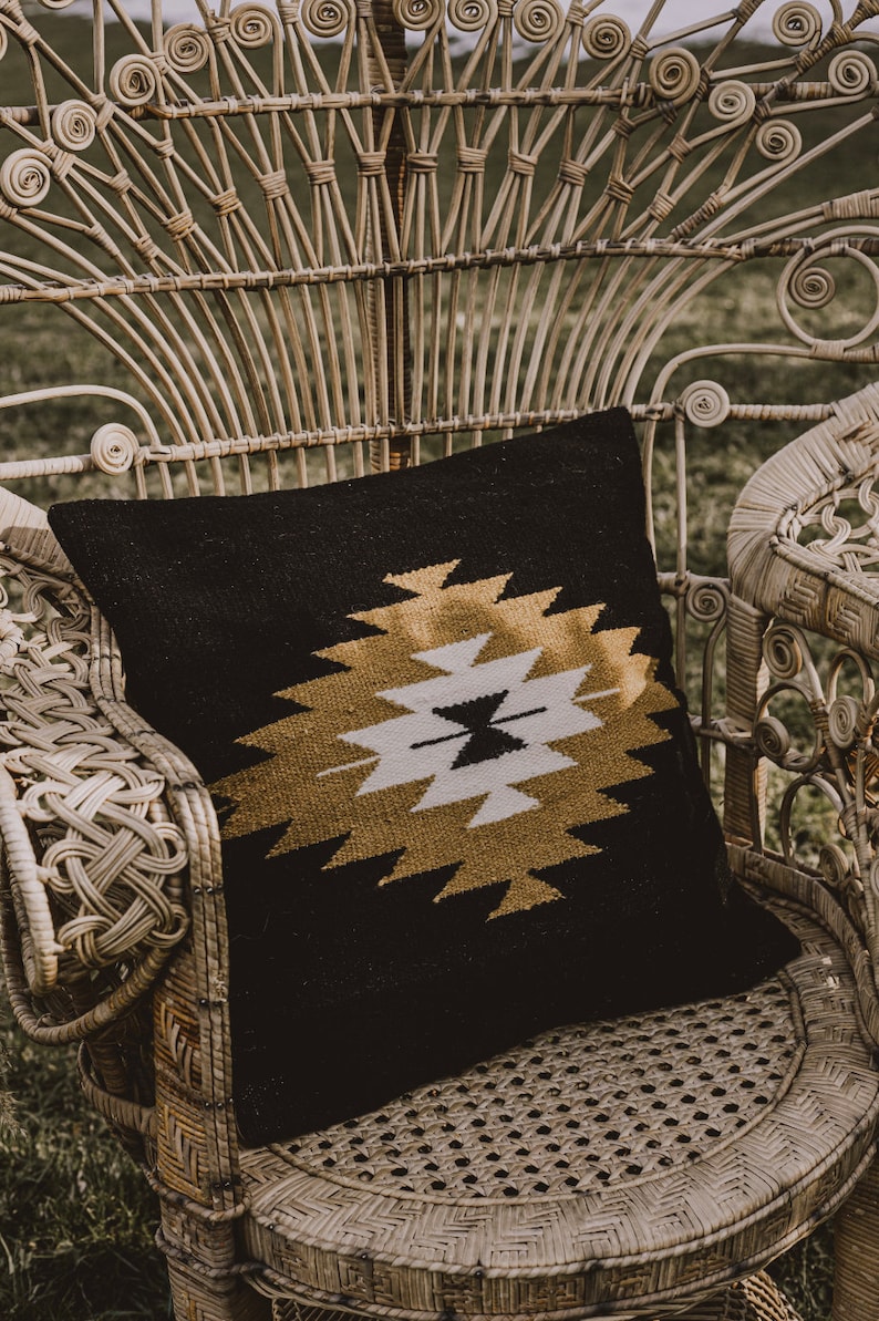 K&ouml;nnte beinhalten: Ein schwarzes Kissen mit goldfarbenem Muster liegt auf einem wei&szlig;en Korbstuhl. Der Stuhl hat ein einzigartiges Design mit einer hohen R&uuml;ckenlehne und geschwungenen Armlehnen.