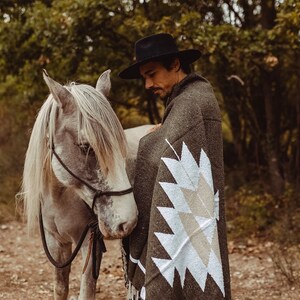 May include: A man wearing a black hat and a green blanket with a white geometric pattern stands next to a white horse. The horse is wearing a black halter and lead rope.