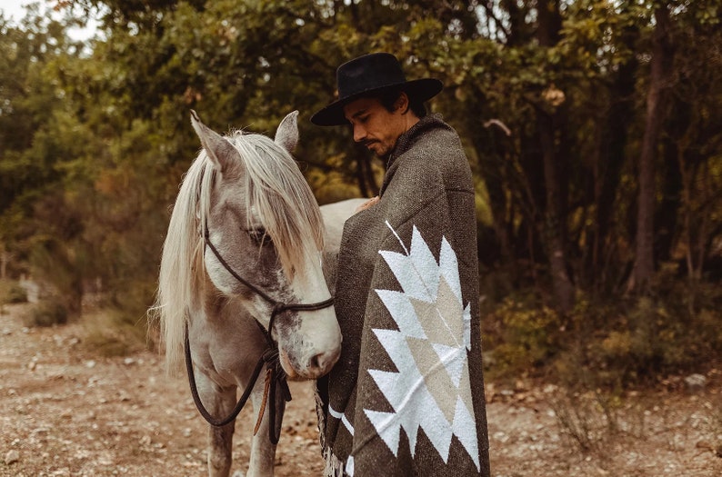 May include: A man wearing a black hat and a green blanket with a white geometric pattern stands next to a white horse. The horse is wearing a black halter and lead rope.